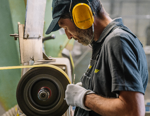 Een APAK-medewerker met gehoorbescherming slijpt een metalen onderdeel.